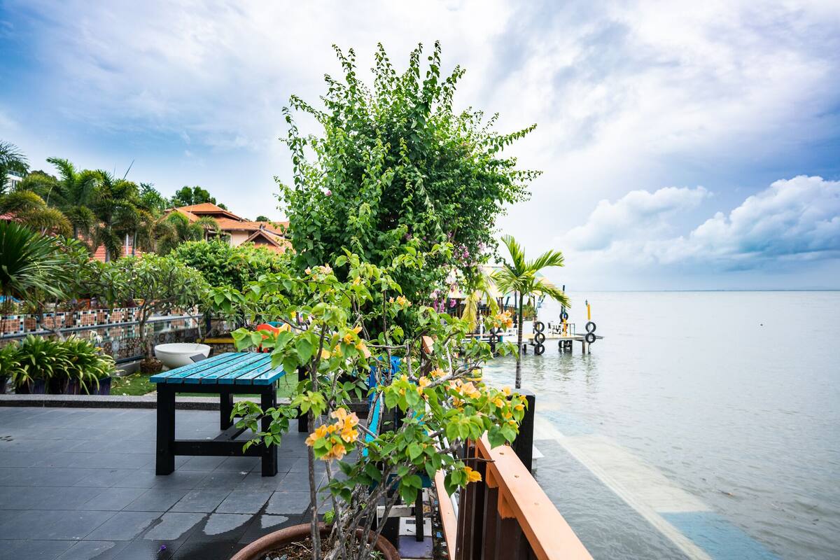 Eine Uferterrasse mit Tisch und Stühlen, umgeben von üppiger Vegetation und Blick auf das Meer.