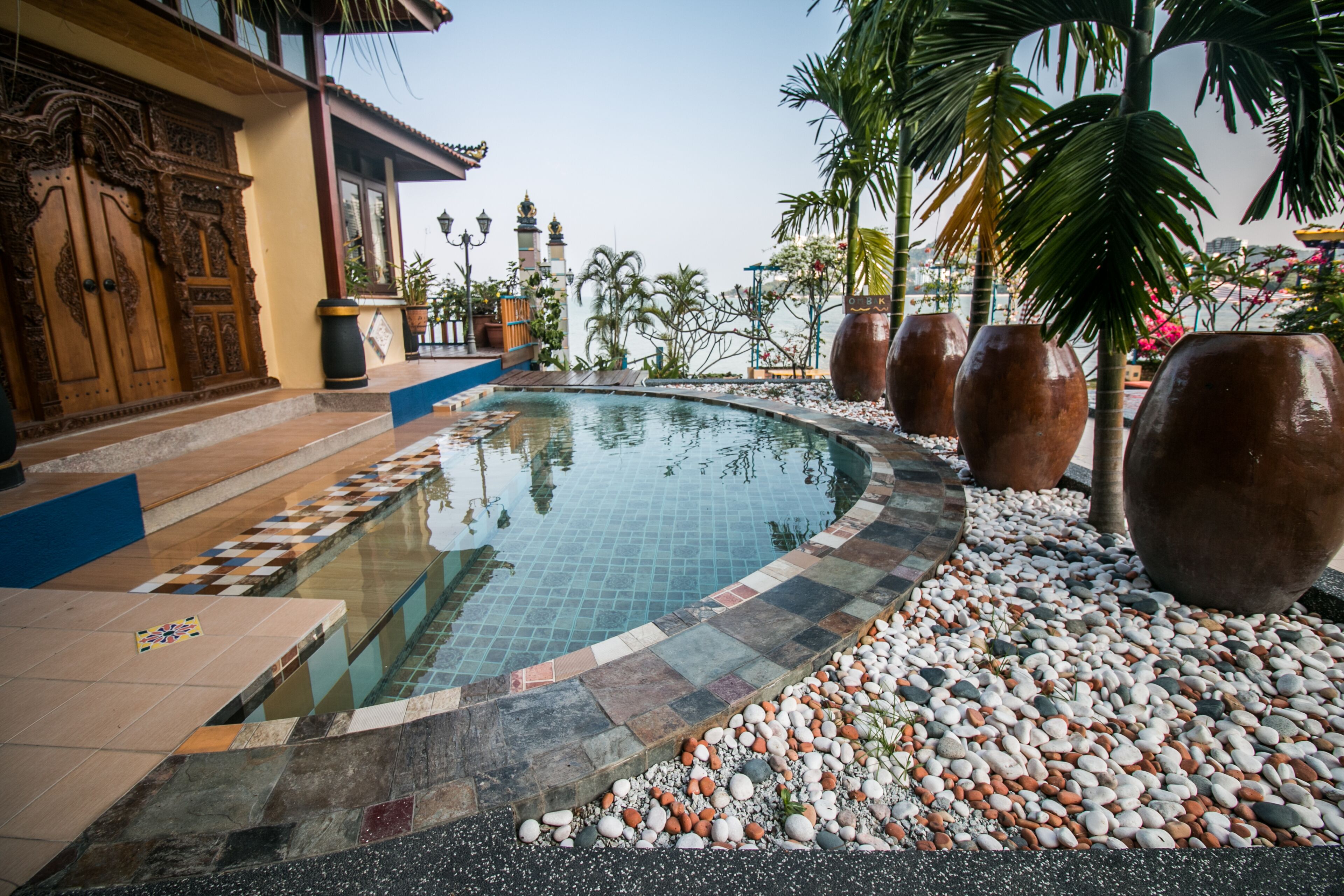 A swimming pool with a tiled edge, surrounded by pebbles and large vases, with a building featuring ornate doors in the background.
