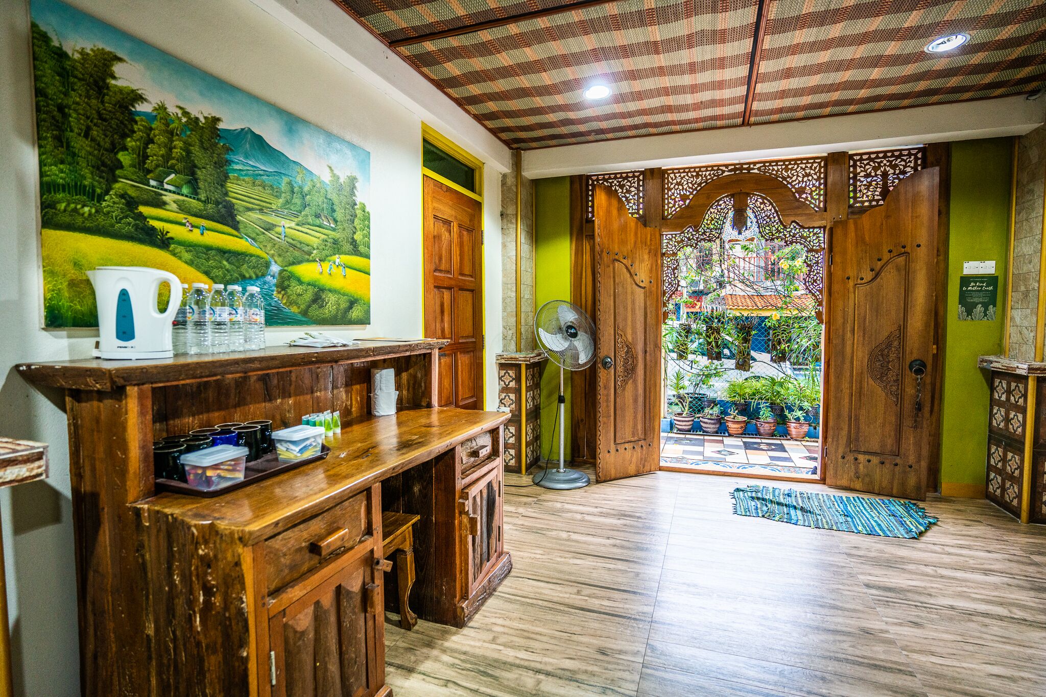 Une pièce avec un comptoir d’accueil en bois, un ventilateur et une vue sur un jardin, visible à travers une porte en bois.