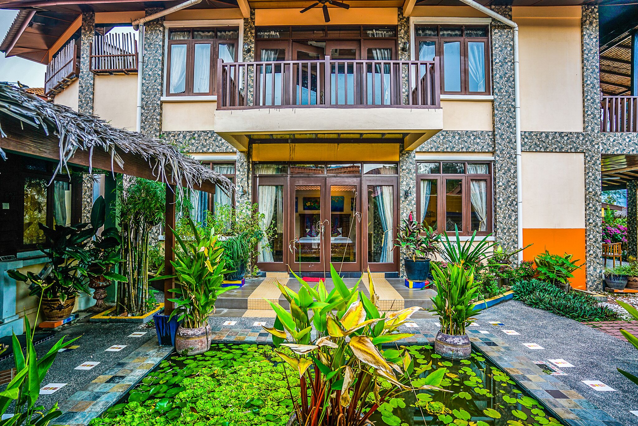 A two-story building with a balcony, a tiled entrance, and a garden with water and plants.