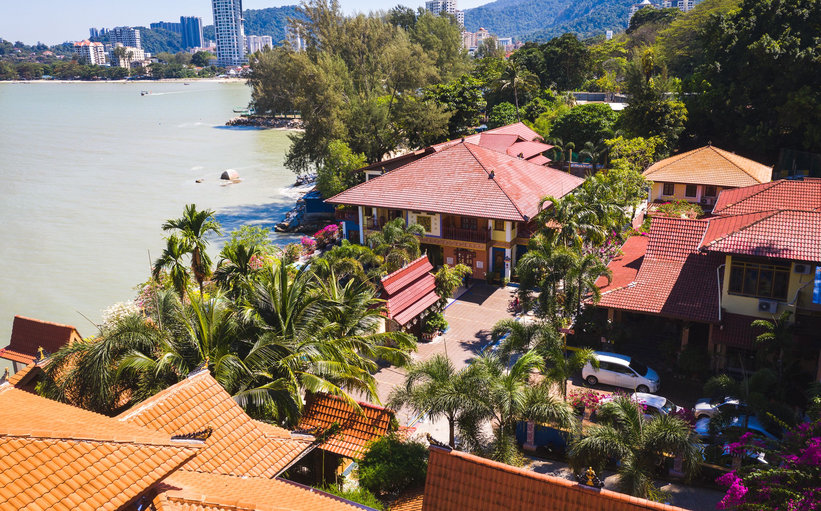 A coastal hotel with red-tiled roofs, surrounded by palm trees and a view of the sea.