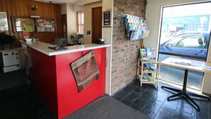A reception area with a red counter, a brick wall, a window with a view of the street, and a small bookshelf. - Midway Pacifica Lodge (Lower Hutt)