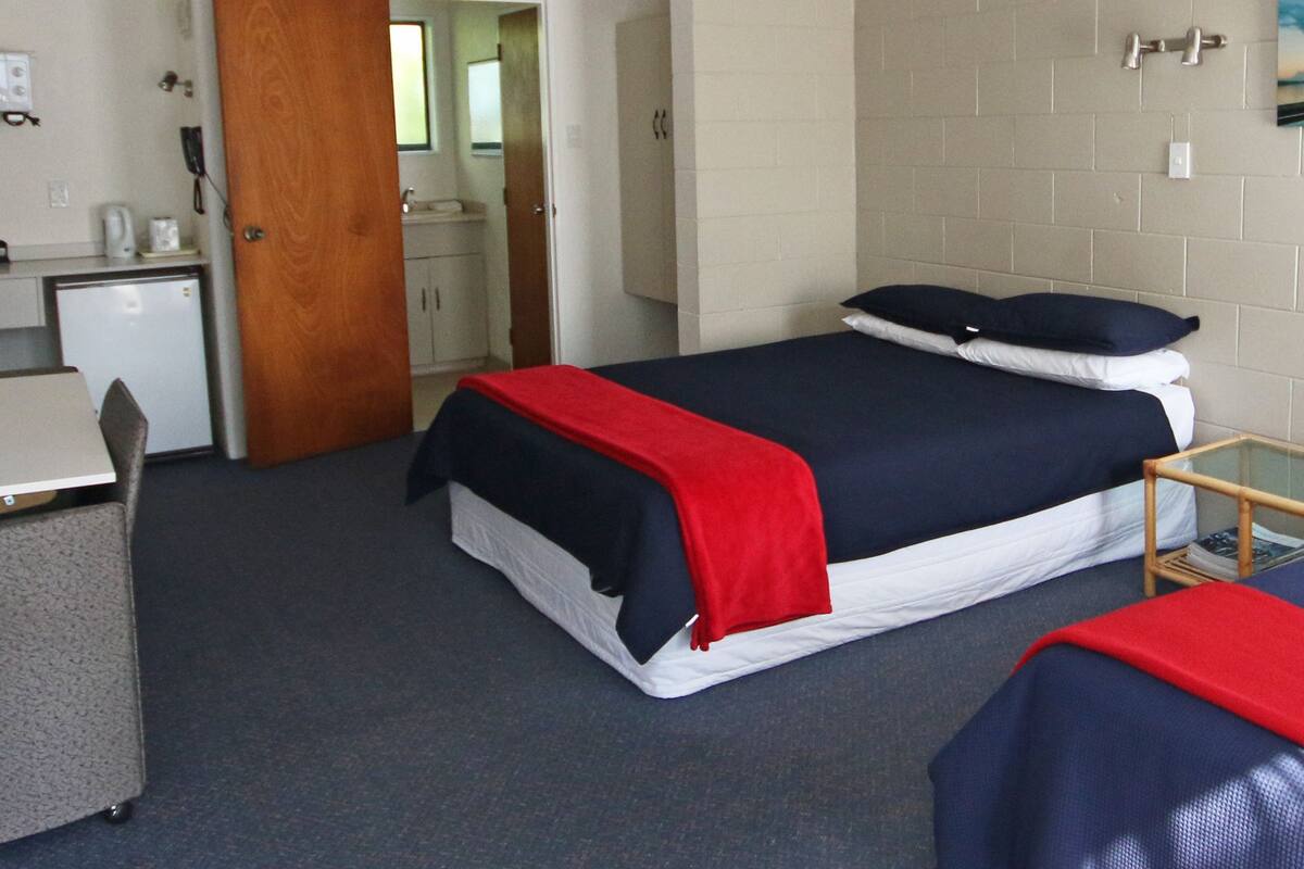 A hotel room with two beds, a desk, a small kitchenette, and a bathroom visible through an open door.