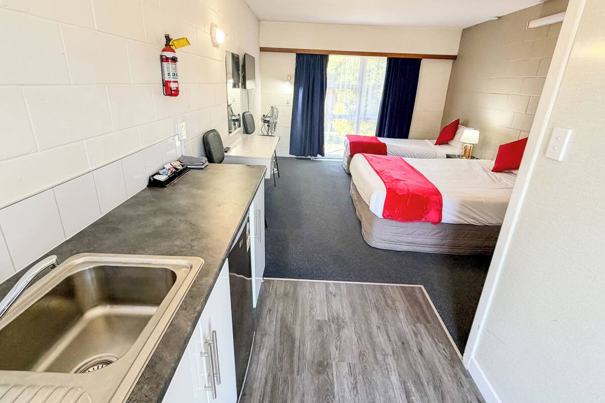 A compact hotel room with a kitchenette, two beds, a small bathroom, and a window with curtains.