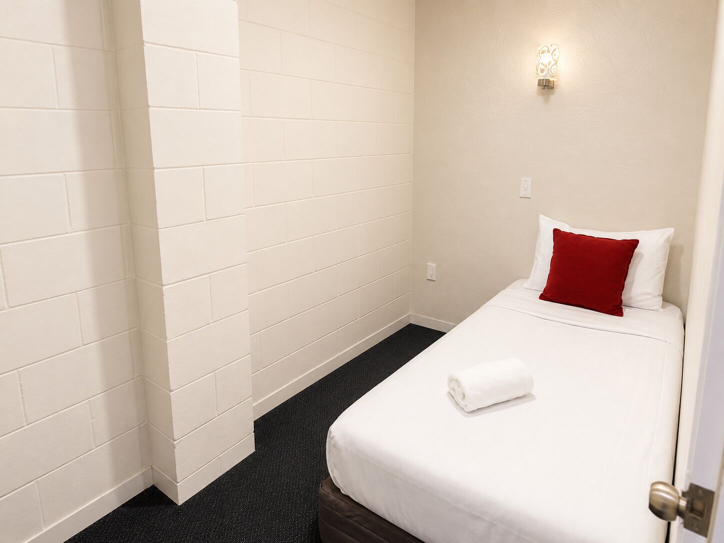 A hotel room with white walls, a single bed with a red pillow, a white towel, and a small wall-mounted light fixture.