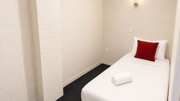 A hotel room with white walls, a single bed with a red pillow, a white towel, and a small wall-mounted light fixture.