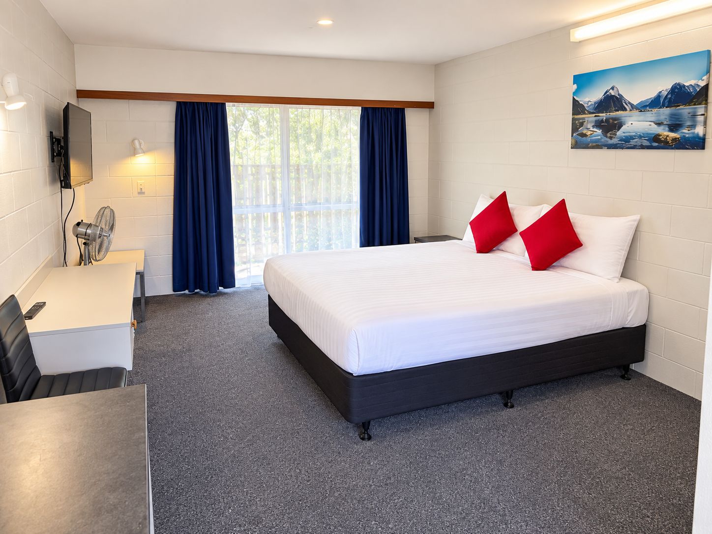 Habitación de hotel con cama, escritorio, silla, televisor montado en la pared y ventana con cortinas.