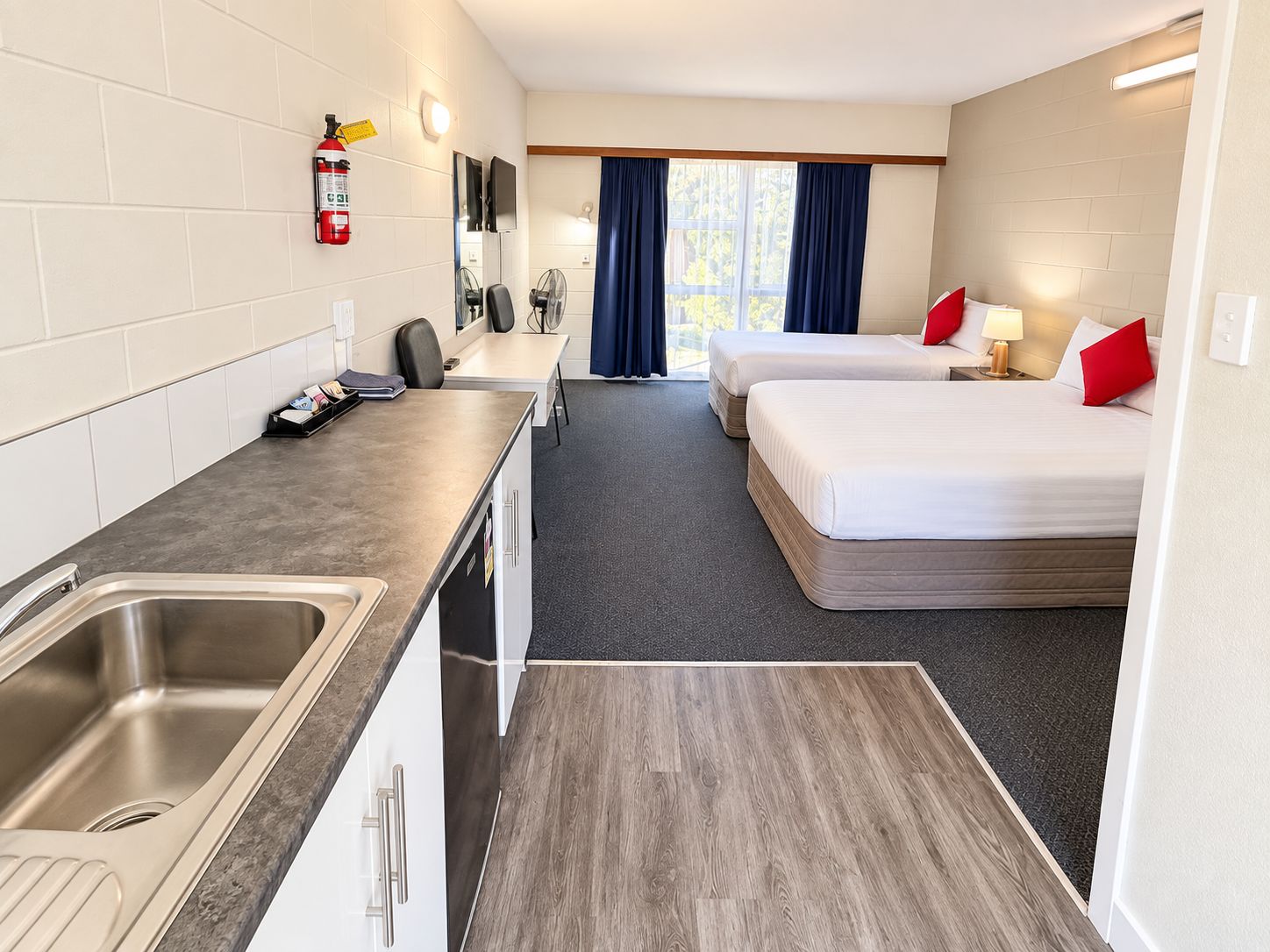 A hotel room with a kitchenette, two beds, a TV, and a window with curtains.