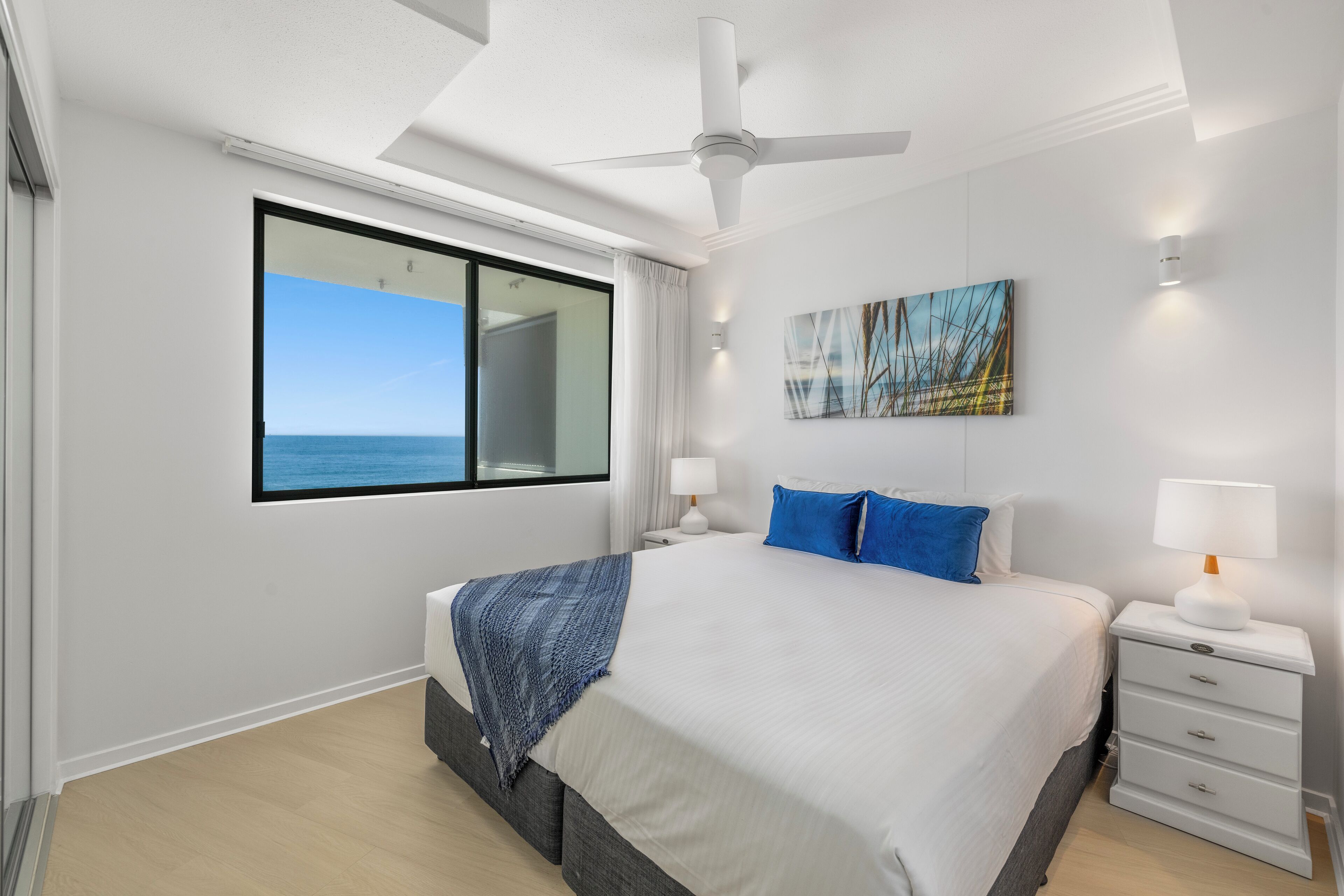 A modern bedroom with a large bed, a ceiling fan, a nightstand with a lamp, a window with a view of the sea, and a painting on the wall.