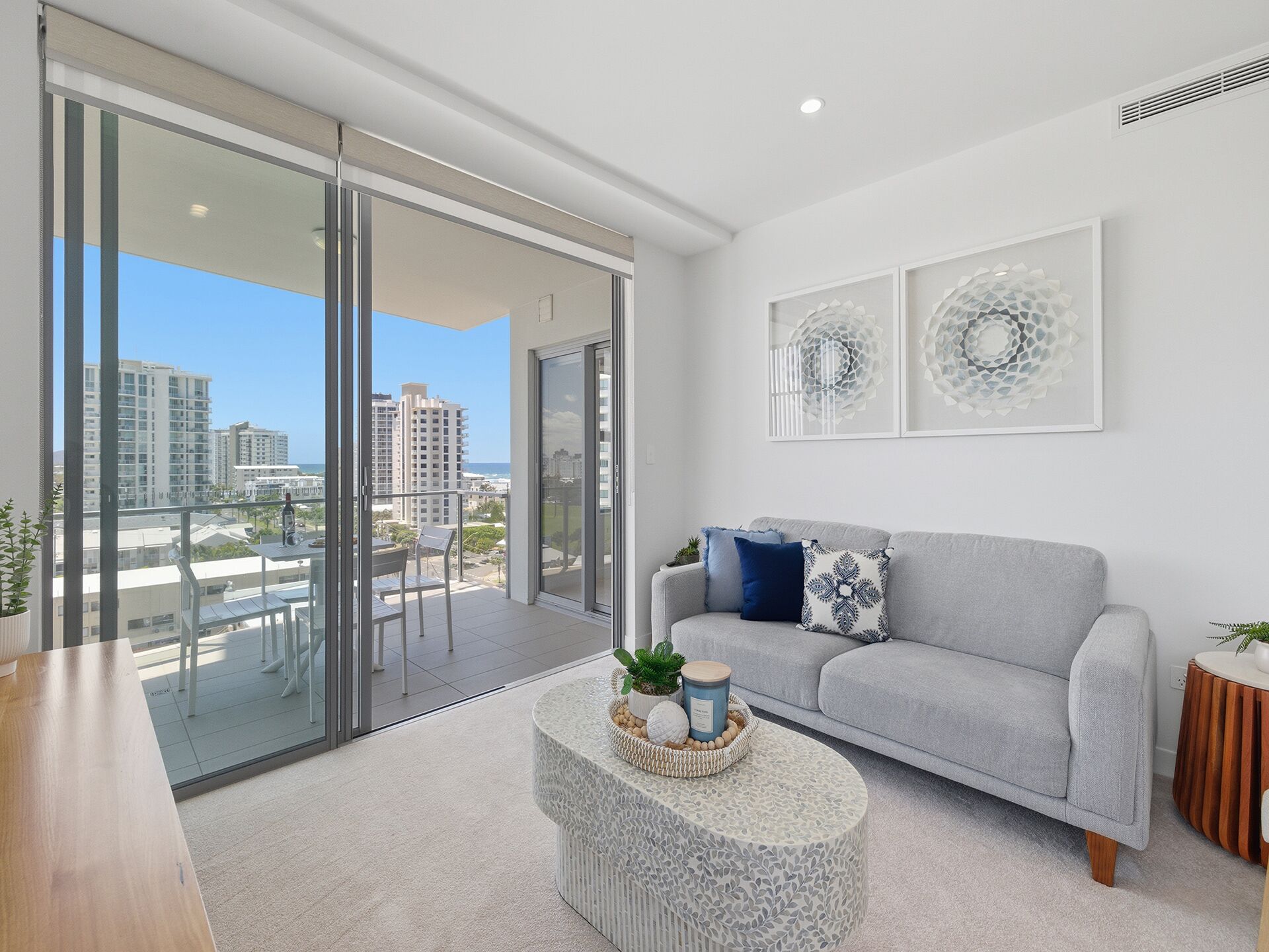 A modern living room with a grey sofa, a round coffee table, and a glass door leading to a balcony with a city view.