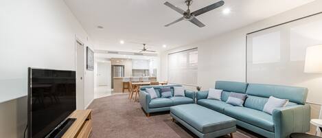 A modern living room with a large flat-screen TV, a blue sectional sofa, and a ceiling fan.