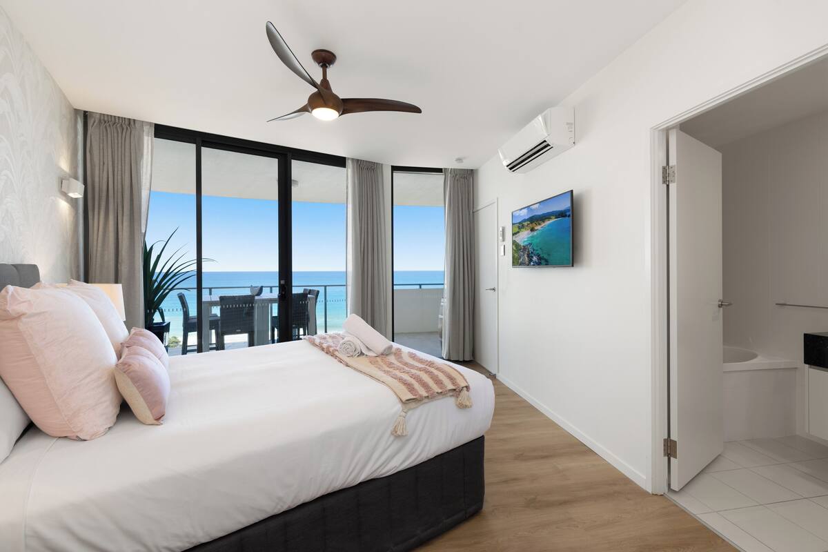 Ein Schlafzimmer mit einem großen Bett, einem Deckenventilator und Blick auf den Ozean durch raumhohe Fenster.