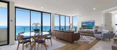 A modern living room with a large window overlooking the sea, a dining area with a table and chairs, and a cozy seating area with a sofa and armchairs.