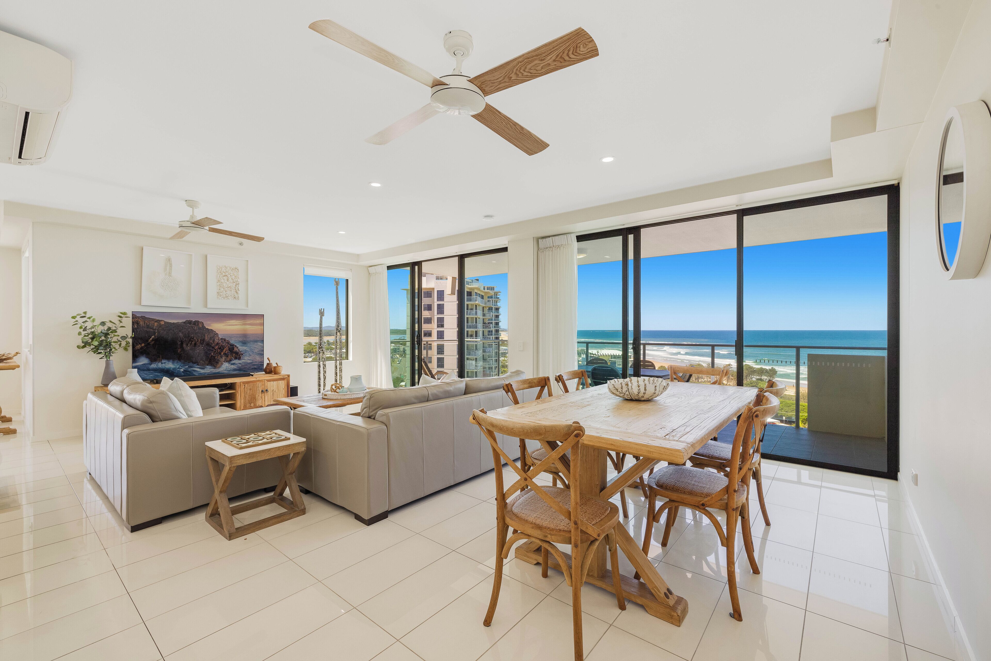 A spacious living room with a large TV, a ceiling fan, and a balcony with a view of the ocean.