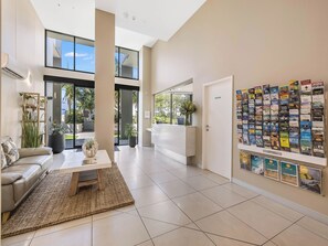 A modern hotel lobby with a reception desk, a seating area with a sofa and coffee table, and a wall displaying various magazines. - Beach on Sixth (Maroochydore)