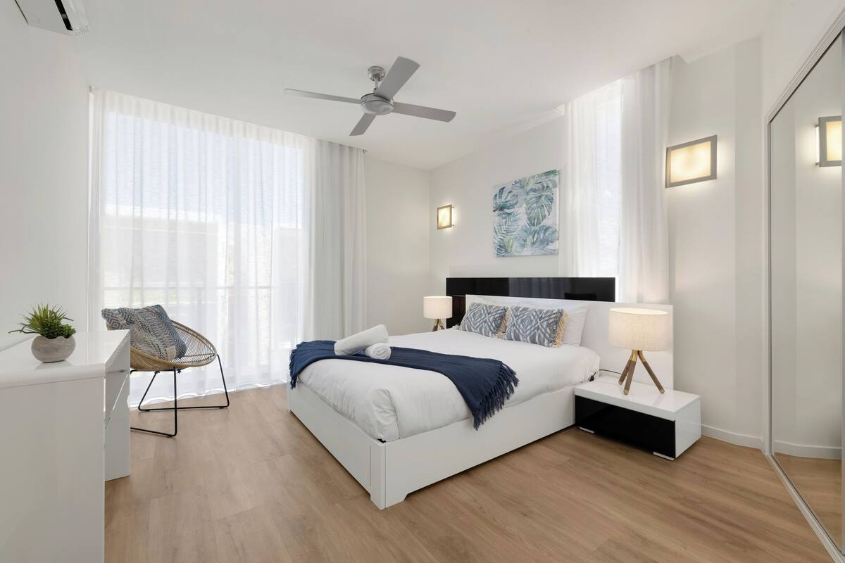 Ein modernes Schlafzimmer mit Bett, Nachttischen, einem Deckenventilator und einem großen Fenster mit durchsichtigen Vorhängen.
