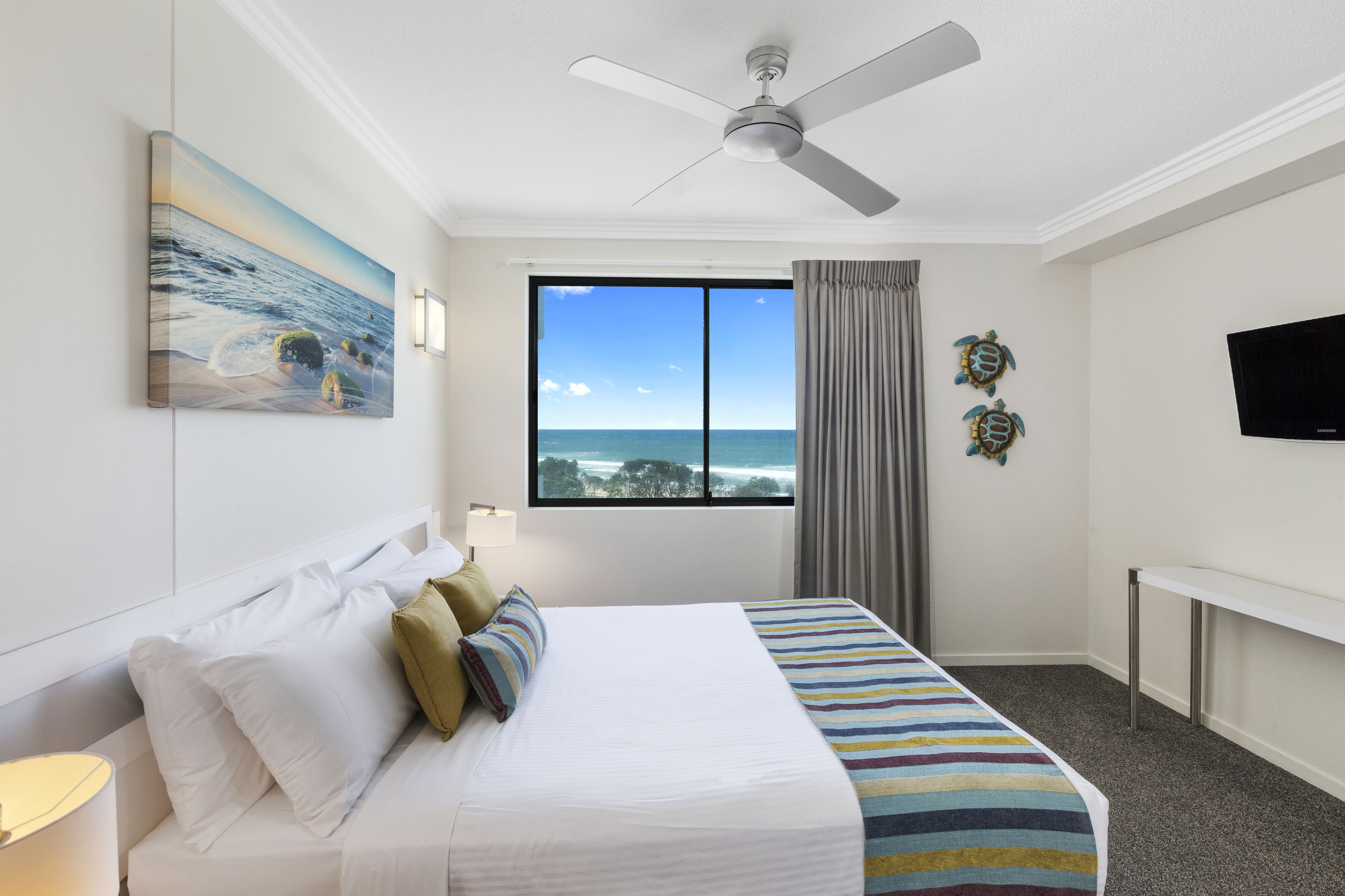 A hotel room with a bed, a ceiling fan, a large window with a view of the ocean, and a flat-screen TV.
