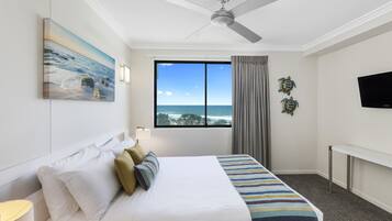 A hotel room with a bed, a ceiling fan, a large window with a view of the ocean, and a flat-screen TV.