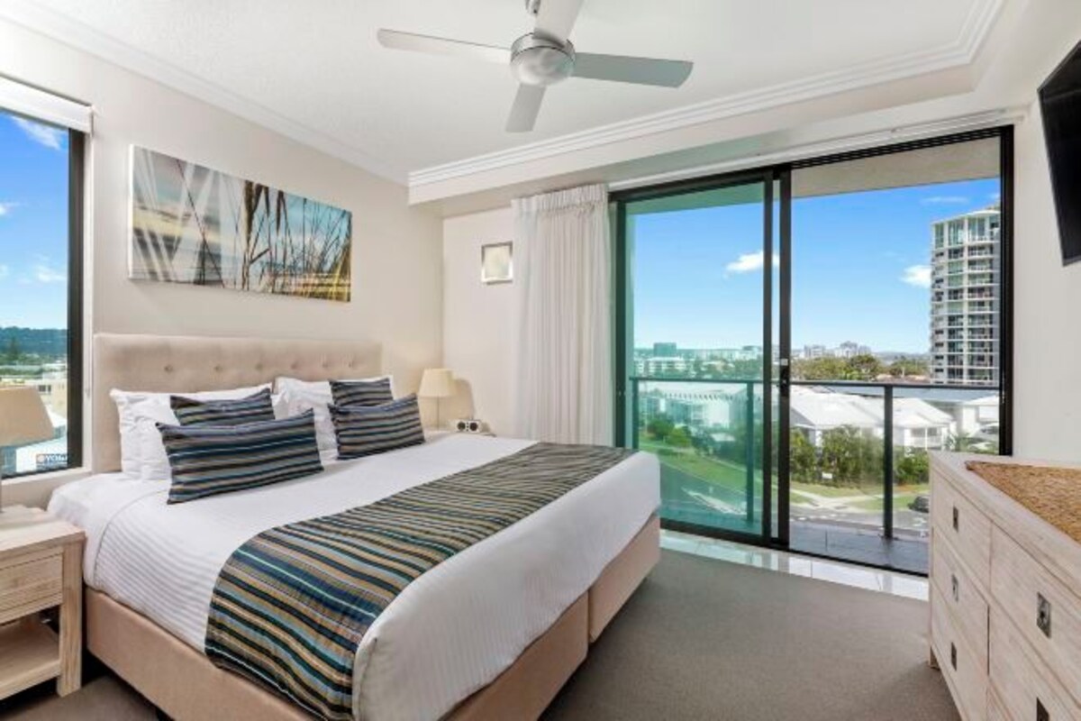 A modern hotel bedroom with a large bed, a ceiling fan, a city view through floor-to-ceiling windows, and a balcony.