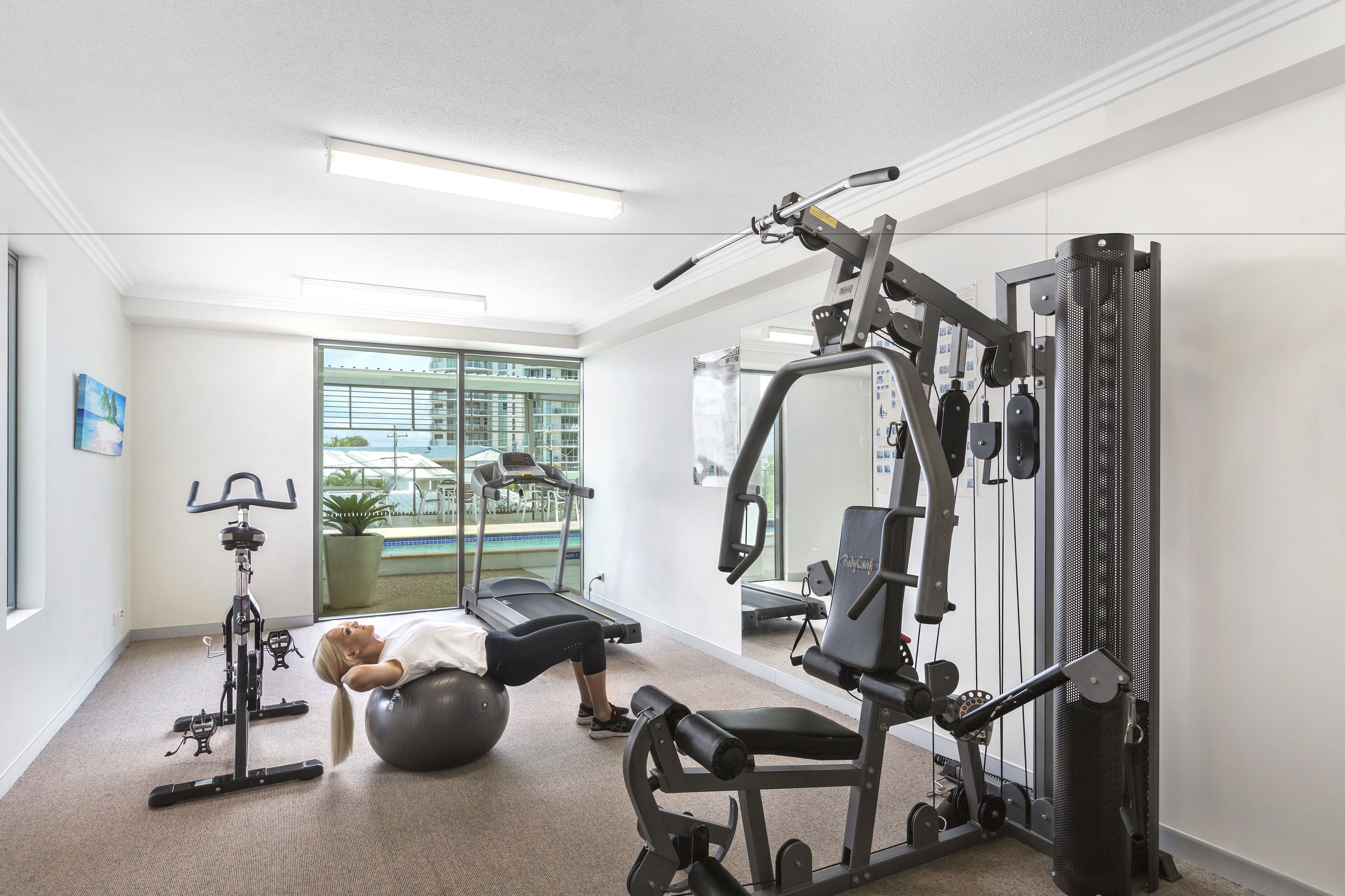 A gym with exercise equipment, a treadmill, and a large window with a view of a building and greenery.