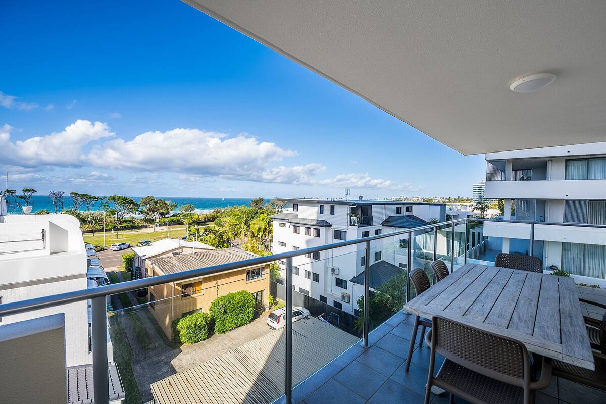 Ein Balkon mit Holztisch und -stühlen, der auf eine Küstenlandschaft mit Gebäuden und einem klaren Himmel blickt.