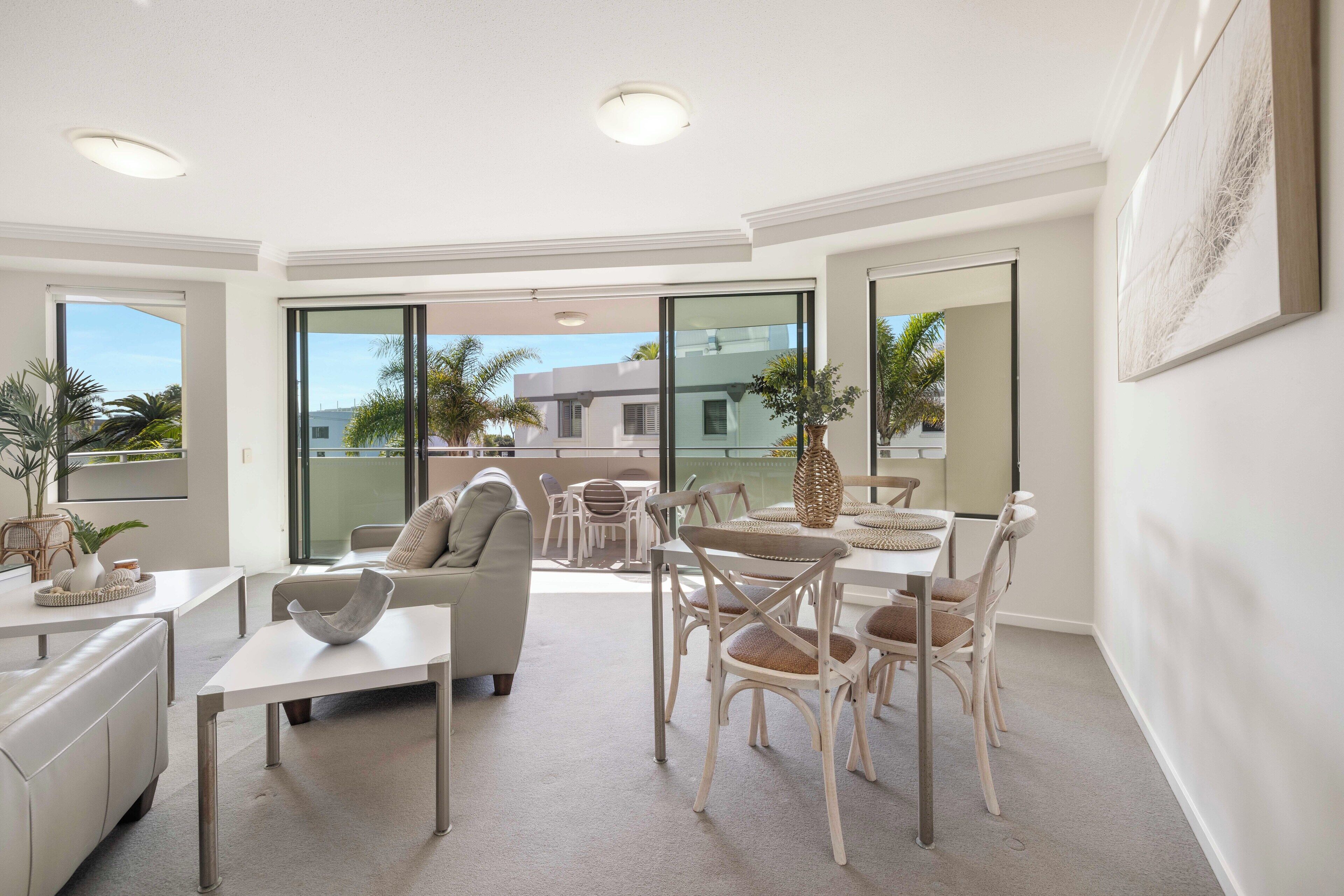 A spacious living area with a dining table, chairs, a sofa, and a coffee table. There are large windows offering a view of palm trees and a building.