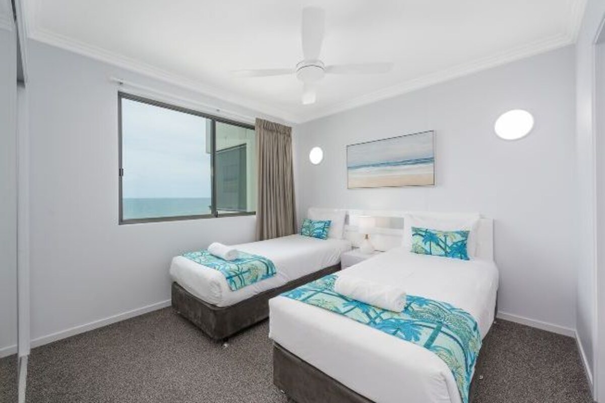 A hotel room with two beds, a ceiling fan, a large window with a view, and a painting on the wall.
