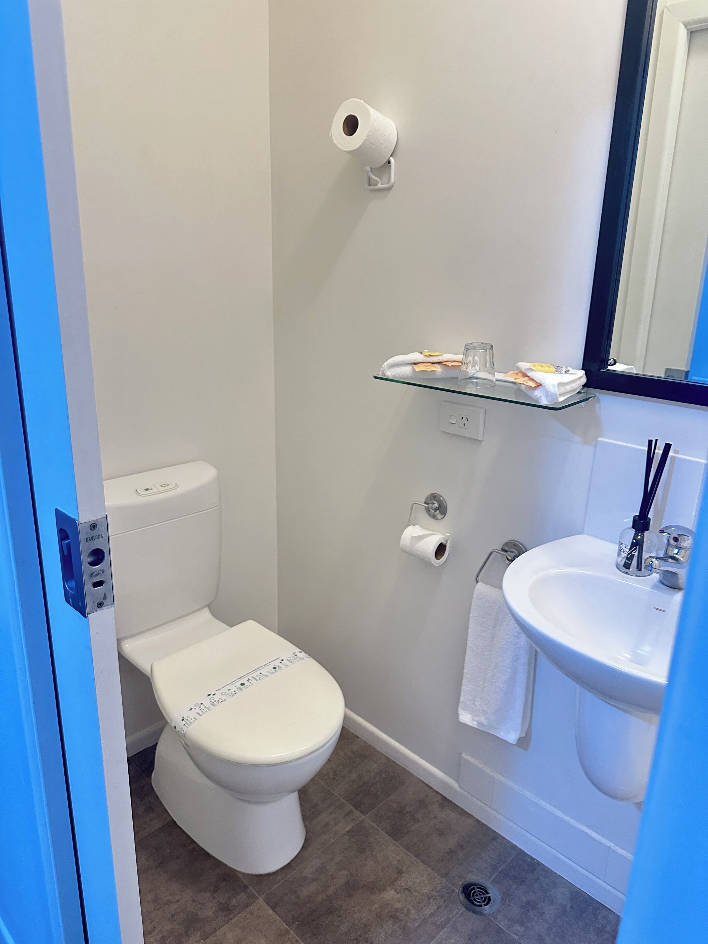A modern bathroom with a toilet, a glass shelf with toiletries, a sink, and a mirror.