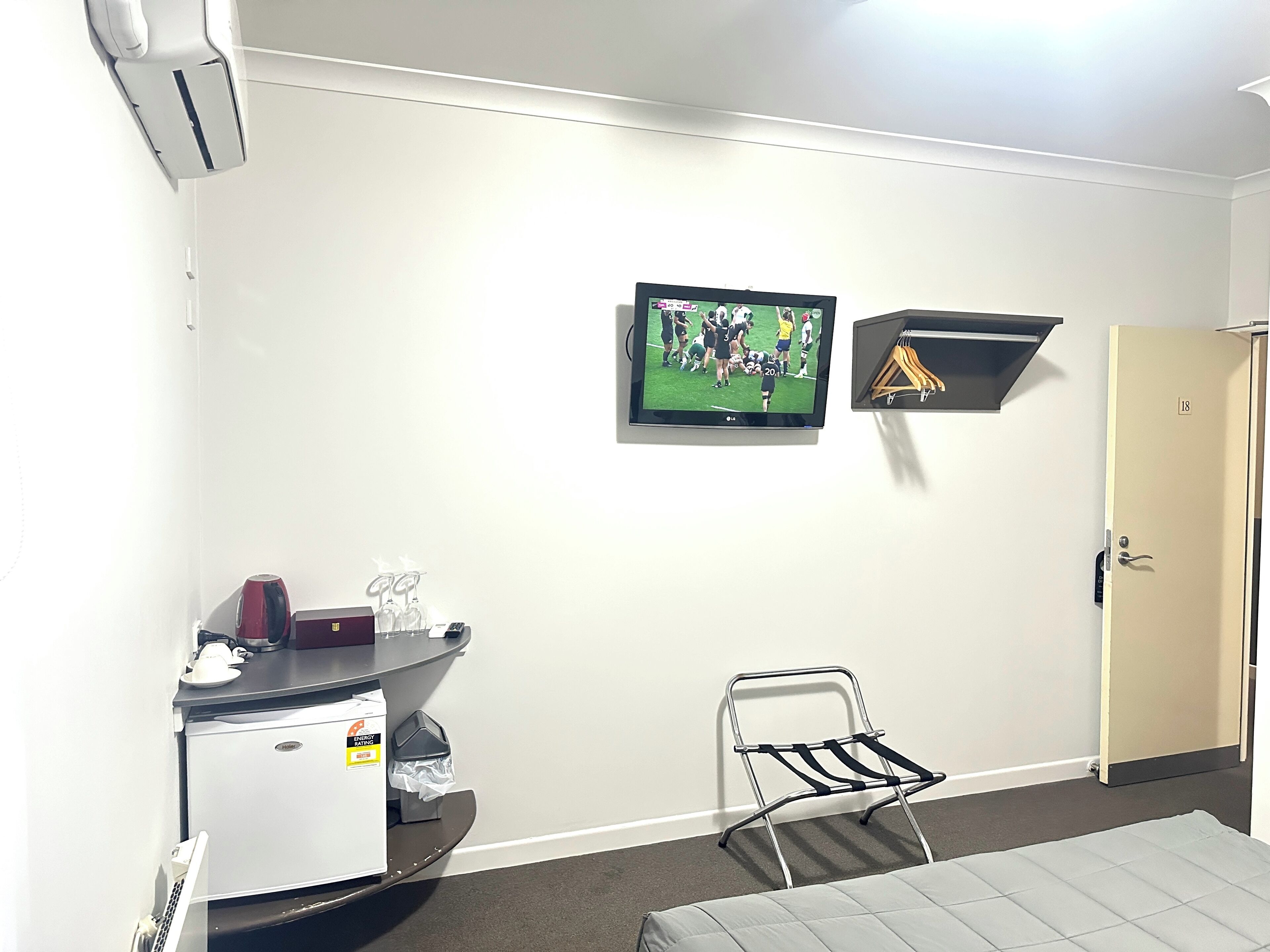 A hotel room with a bed, a desk with a computer, a chair, a TV mounted on the wall, and a door with a number.
