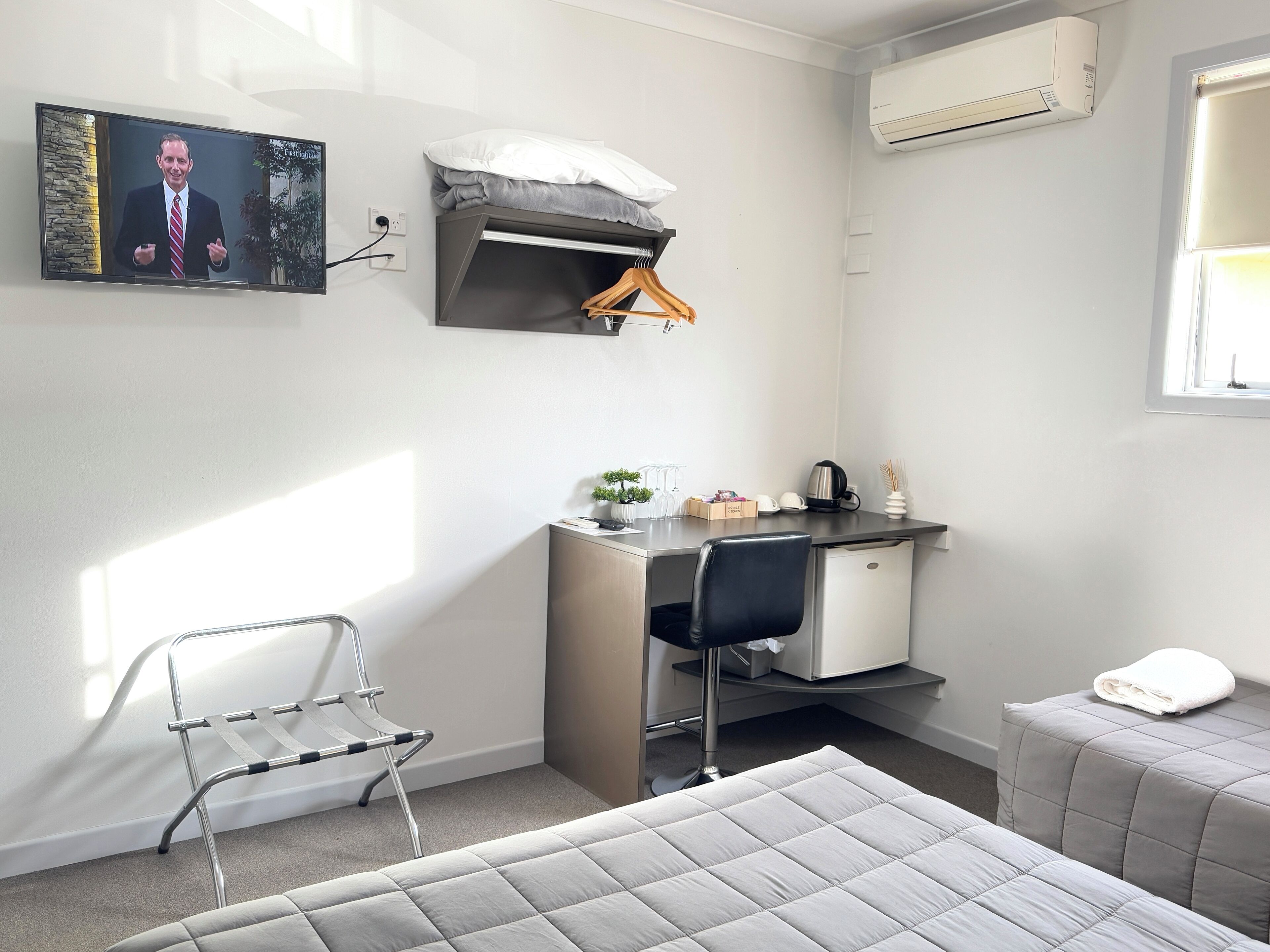 A modern, well-lit room with a bed, a desk with a chair, a hanging clothes rack, an air conditioning unit, and a window.