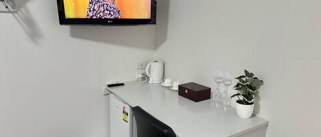 Quarto de hotel com escrivaninha branca, televisão na parede, planta em vaso, cafeteira e uma caixa.