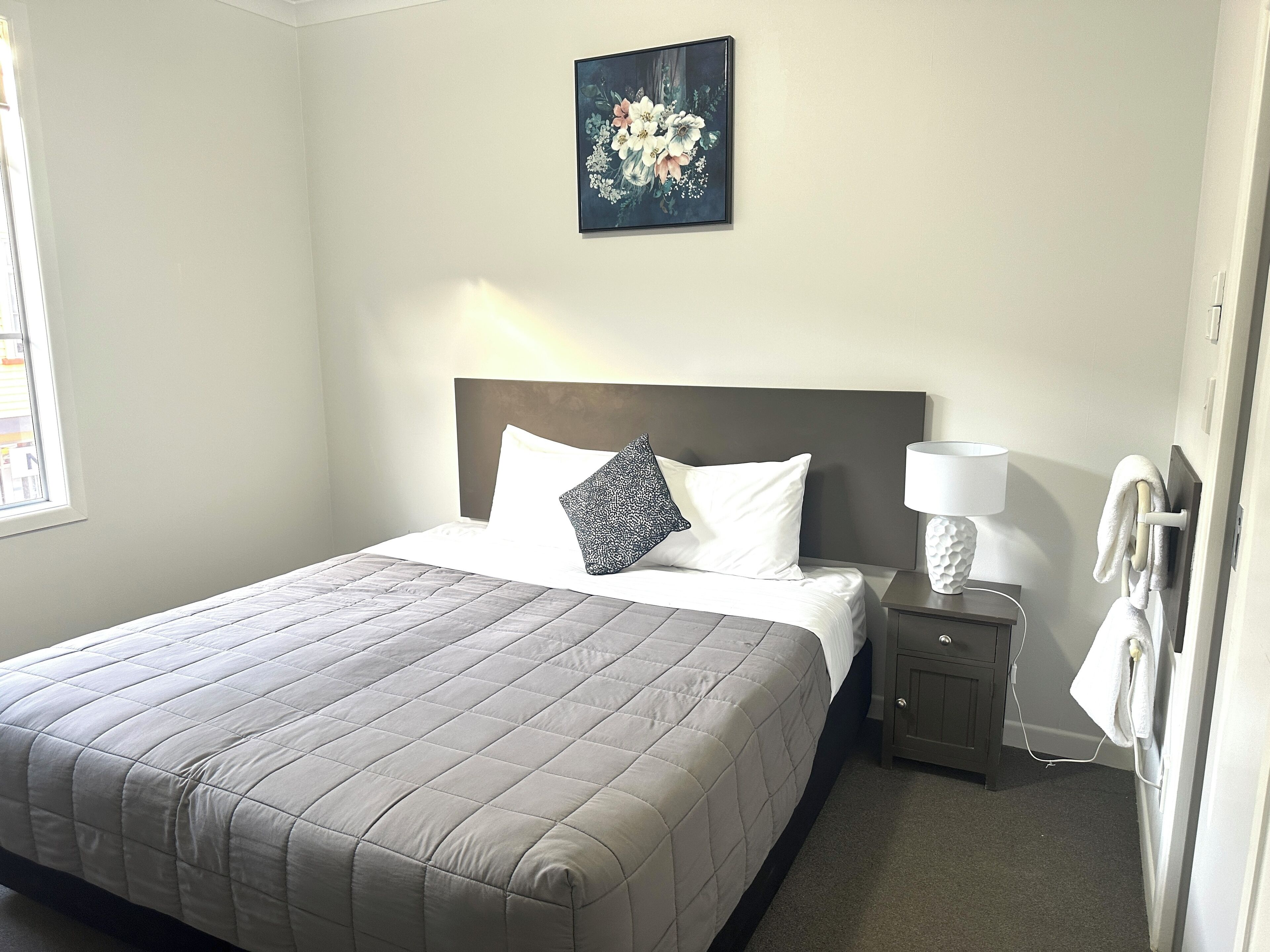 A neatly made bed with a quilted bedspread, a pillow, and a bedside table with a lamp and a towel hanging on the wall.