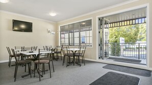 A dining area with a large table and chairs, a flat-screen TV on the wall, and a view of the outdoors through large windows. - No1 Motels On Victoria (Cambridge)