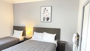 A hotel room with two beds, a nightstand with a lamp, a towel rack, and a framed artwork on the wall.