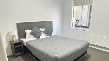 Ein ordentlich bezogenes Bett mit einer grauen Decke, ein Nachttisch mit einer Lampe und ein Fenster mit Blick nach draußen.