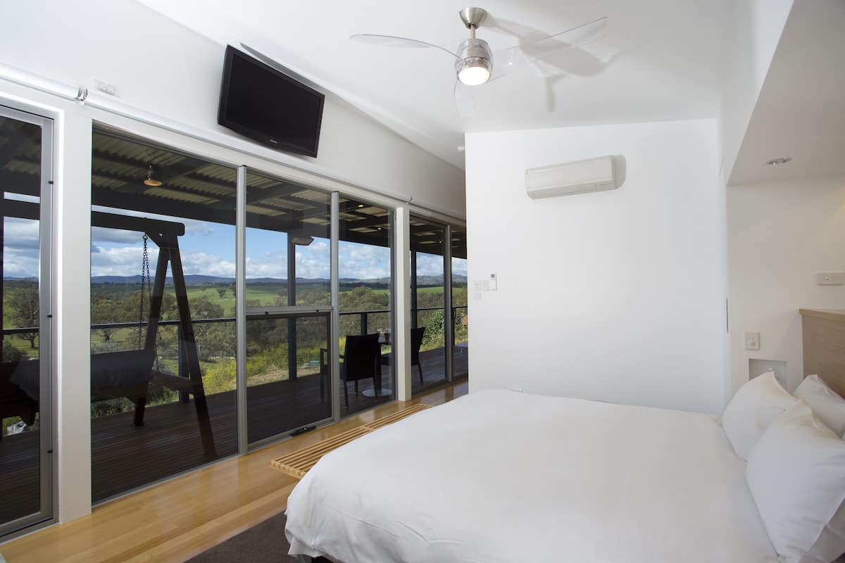 A bedroom with a bed, a ceiling fan, a TV, and a large window with a view of the outdoors.