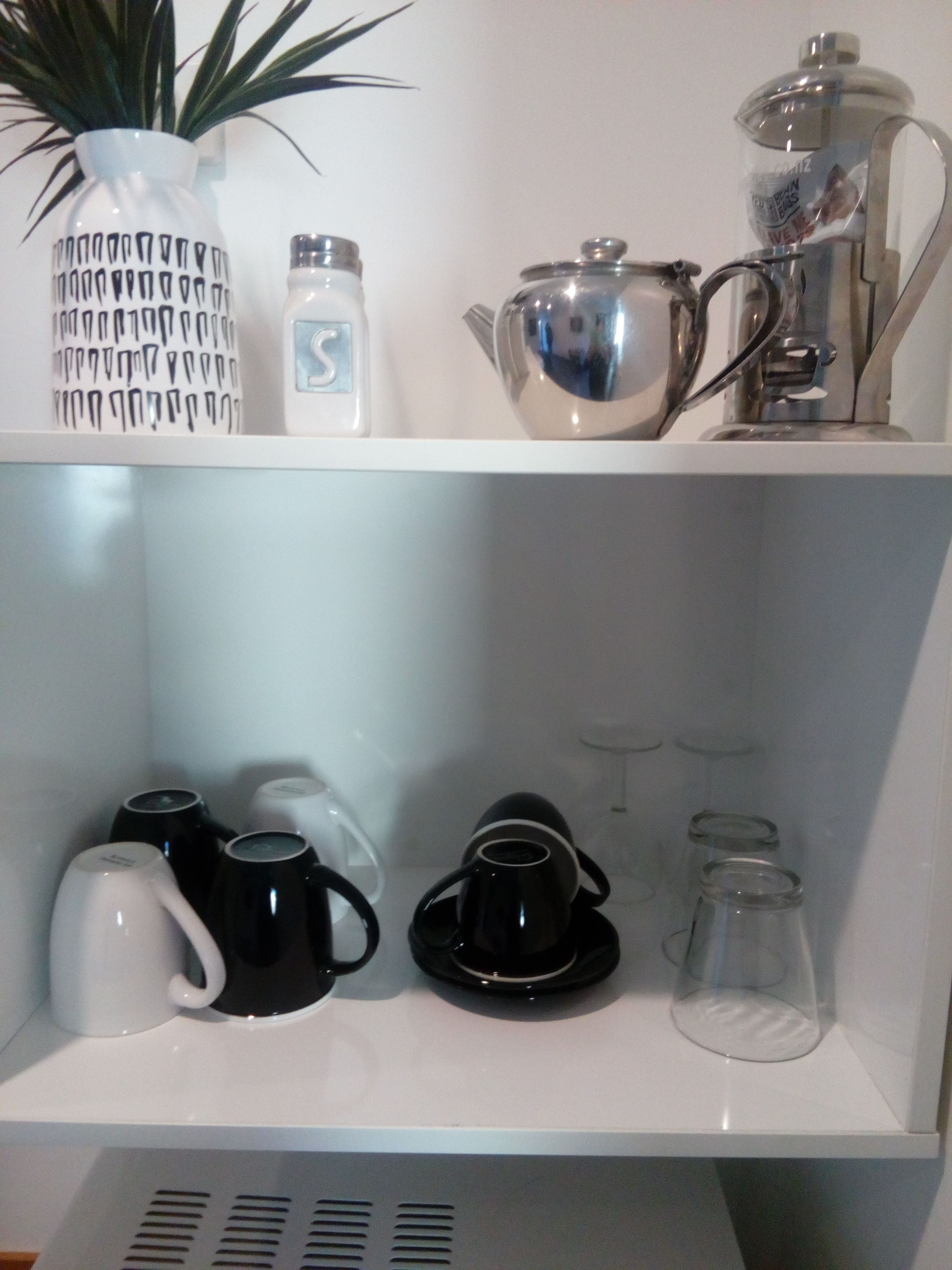 A kitchen shelf with a coffee maker, a teapot, a vase with a plant, and various cups and mugs.