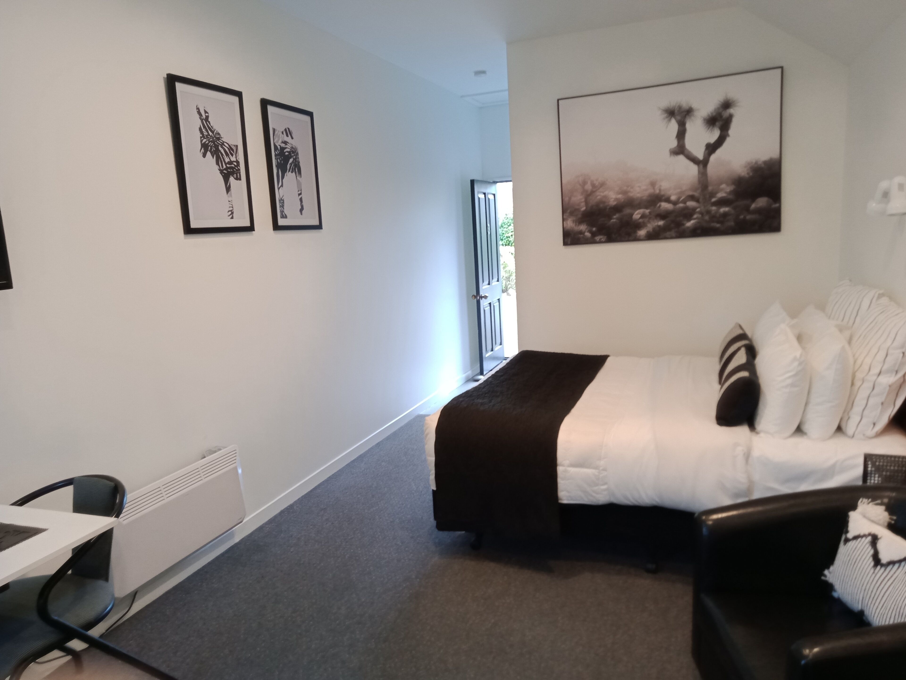 A hotel room with a bed, a desk, and chairs. There are two framed pictures on the wall and a large painting above the bed.