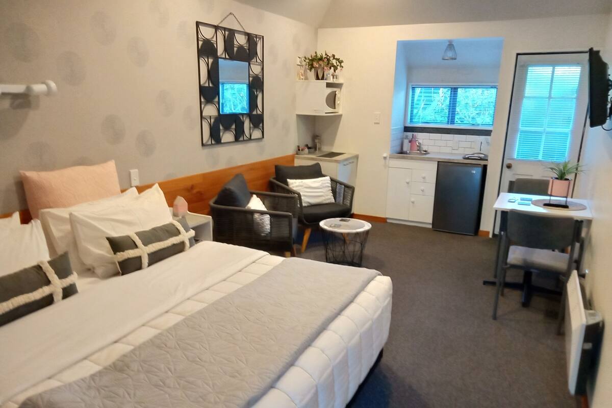 A hotel room with a large bed, a desk, a chair, a small table, a microwave, a sink, and a window with blinds.