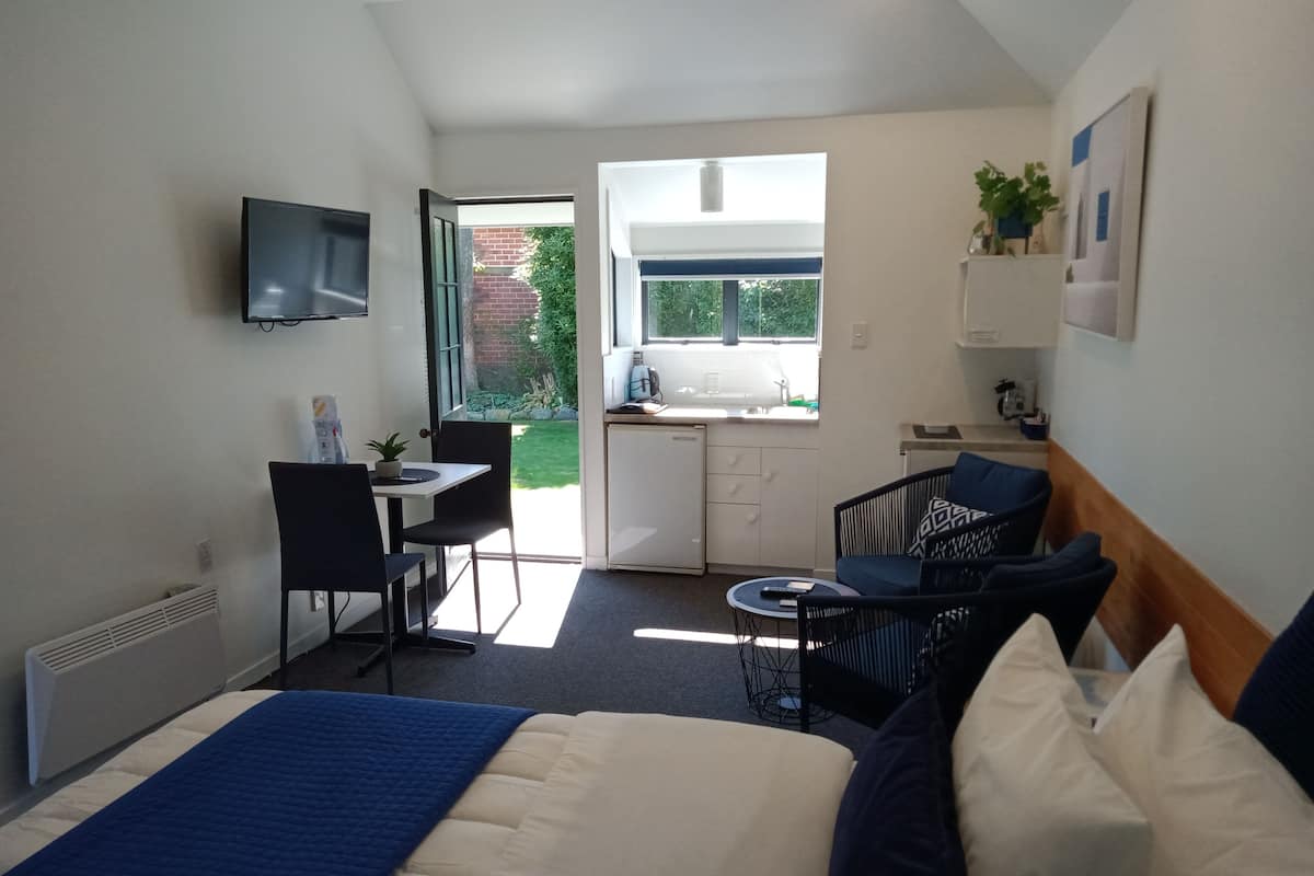 A compact hotel room with a bed, a desk, a chair, a small table, a TV, and a kitchenette.