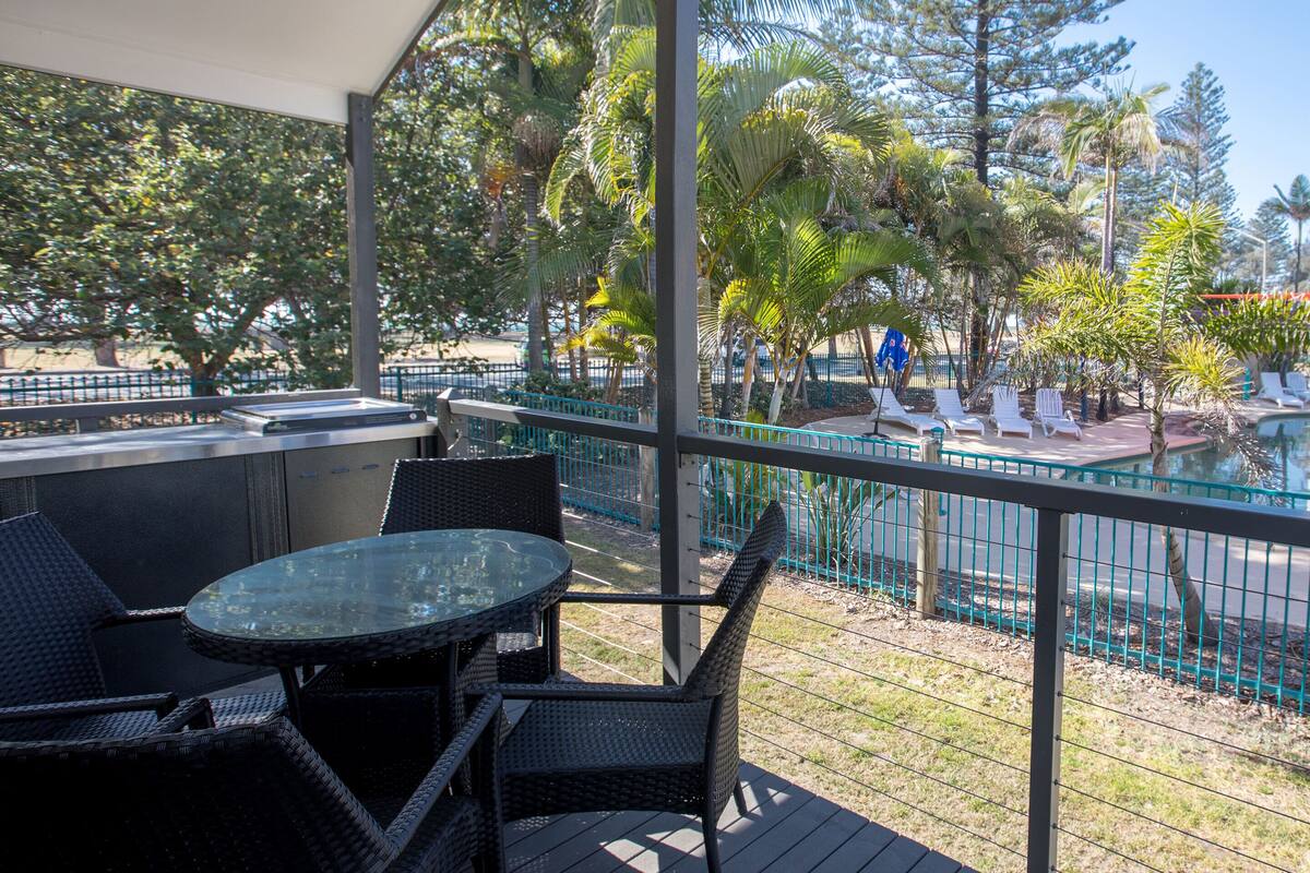 A covered outdoor area with a glass table and chairs, overlooking a pool area with lounge chairs and palm trees.