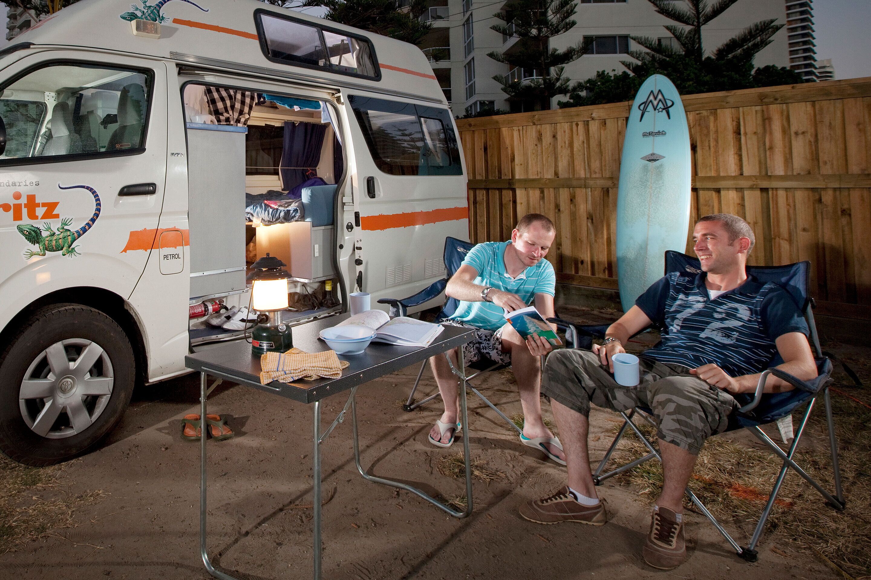 Two people sitting at an outdoor table with a van and a surfboard in the background.