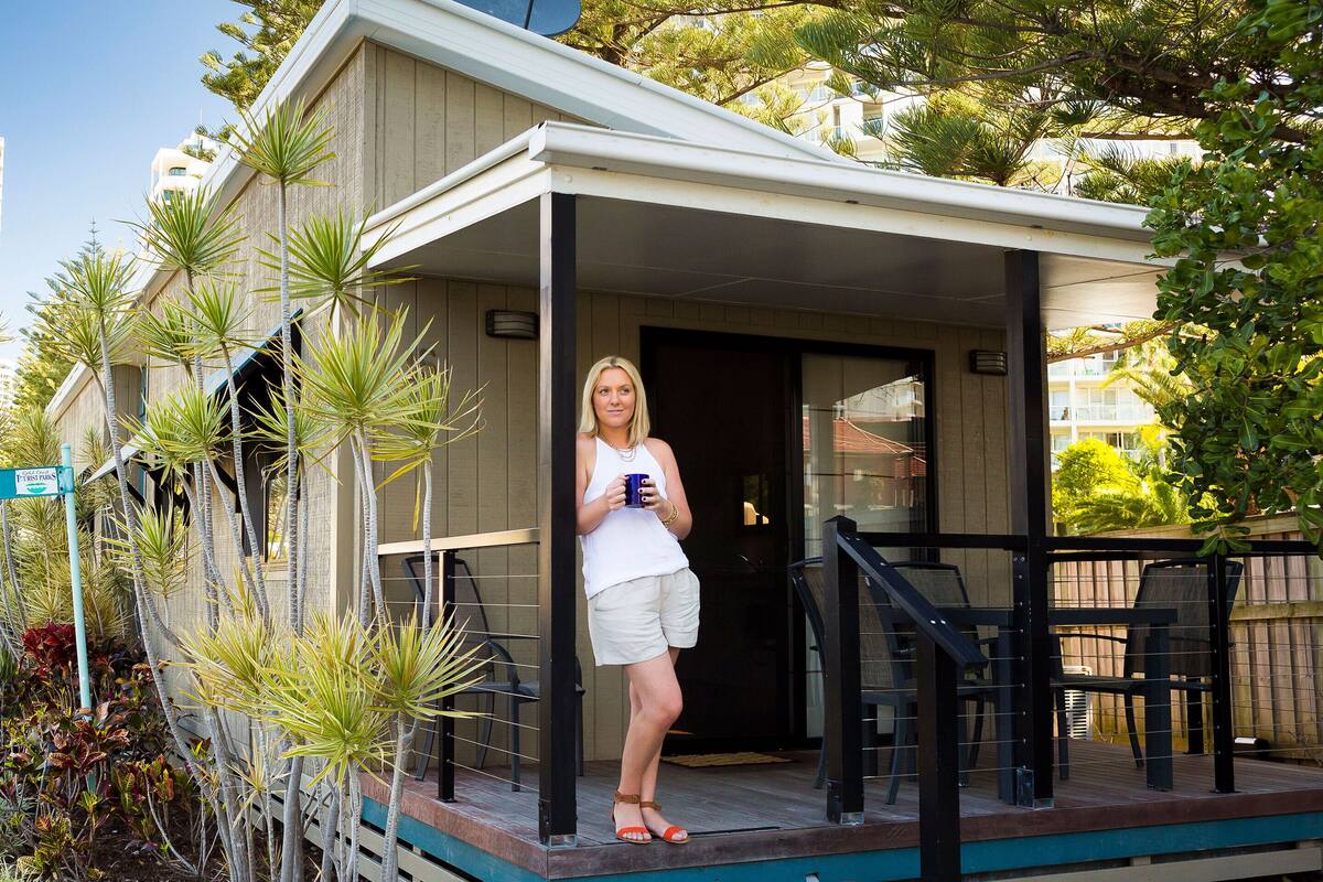 A person standing on a covered porch with a pool, surrounded by palm trees and a wooden fence.