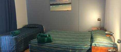 A room with two single beds, a ceiling fan, a painting of a beach, a desk with a lamp, and a wooden floor.