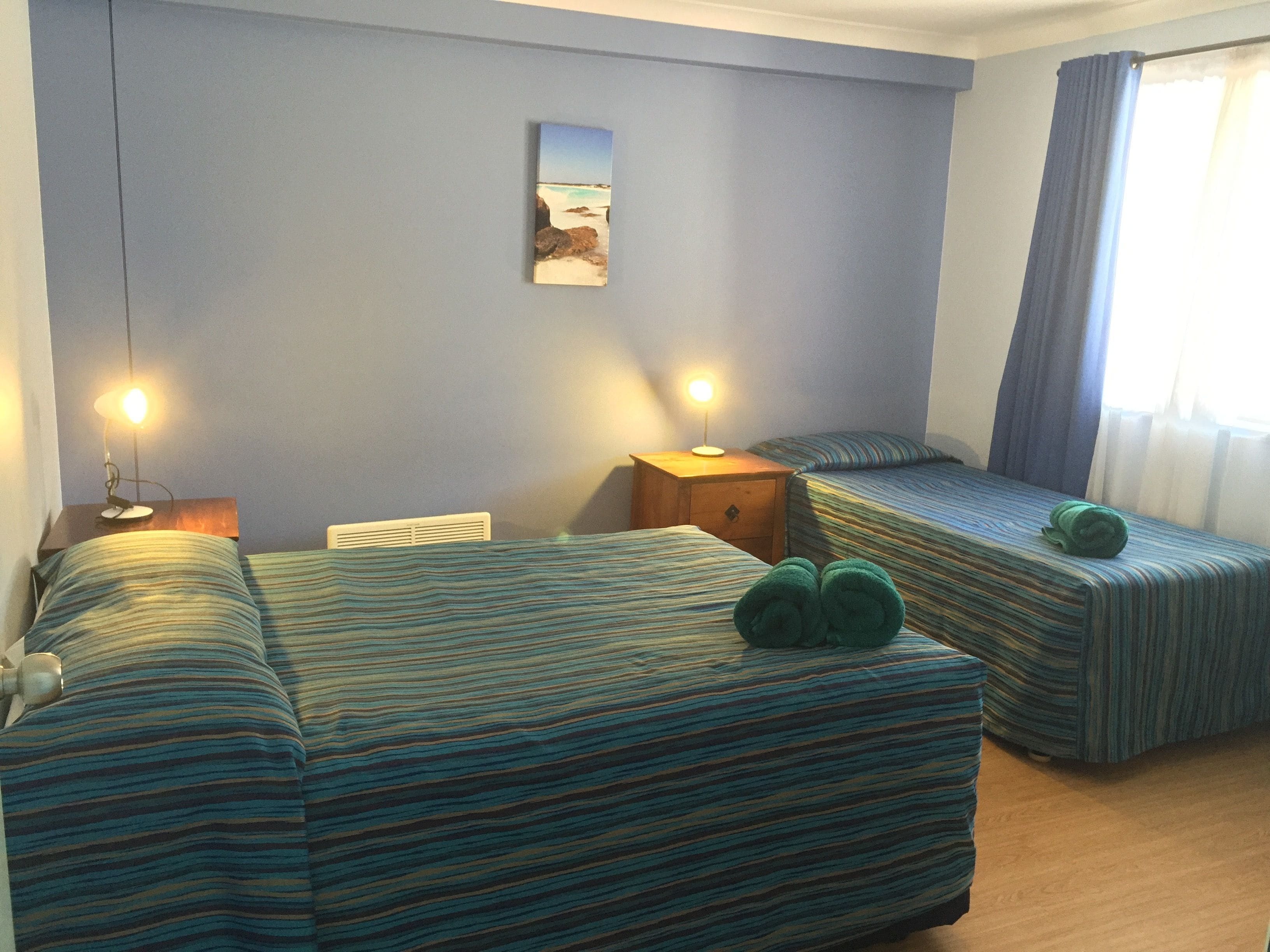 Una habitación con dos camas, una mesita de noche con lámpara, un cuadro en la pared y una ventana con cortinas.