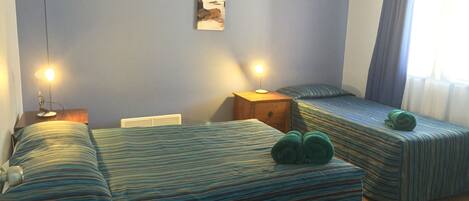 Una habitación con dos camas, una mesita de noche con lámpara, un cuadro en la pared y una ventana con cortinas.