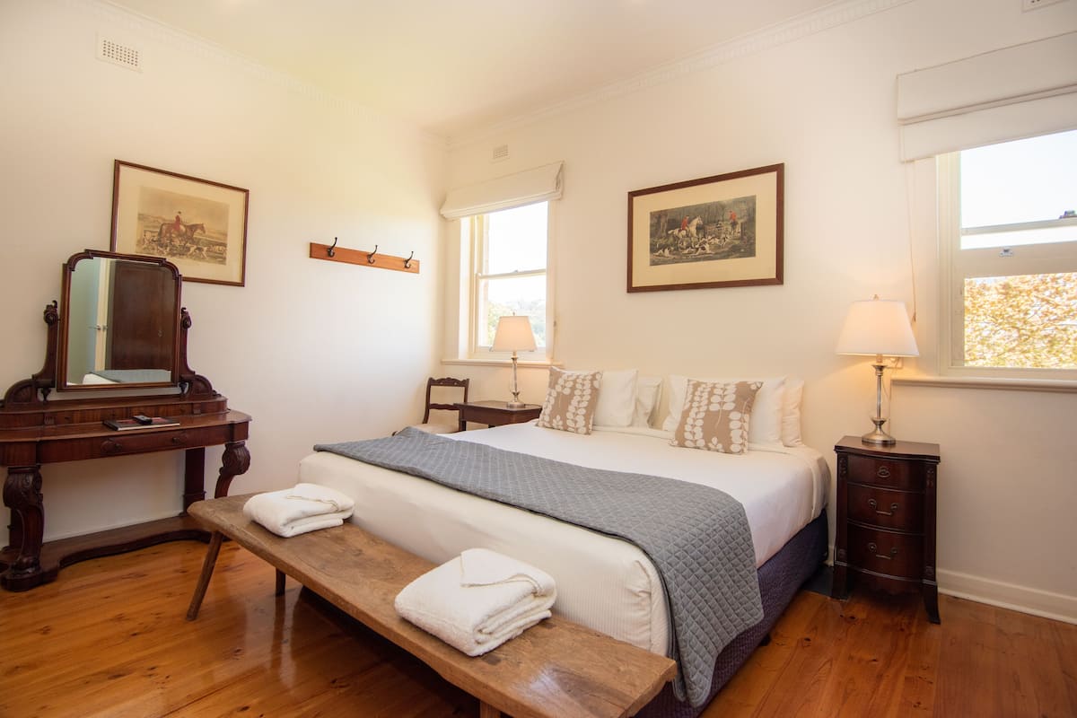 Un dormitorio con una cama, mesitas de noche, un banco, un aparador y dos cuadros en la pared.