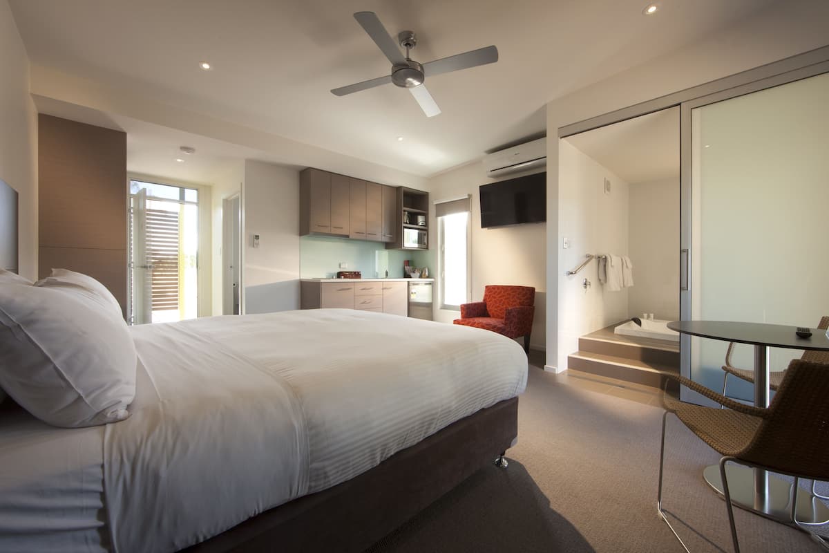 Un dormitorio moderno con una cama grande, ventilador de techo, baño con paredes de vidrio, zona de cocina y mesa de comedor con sillas.