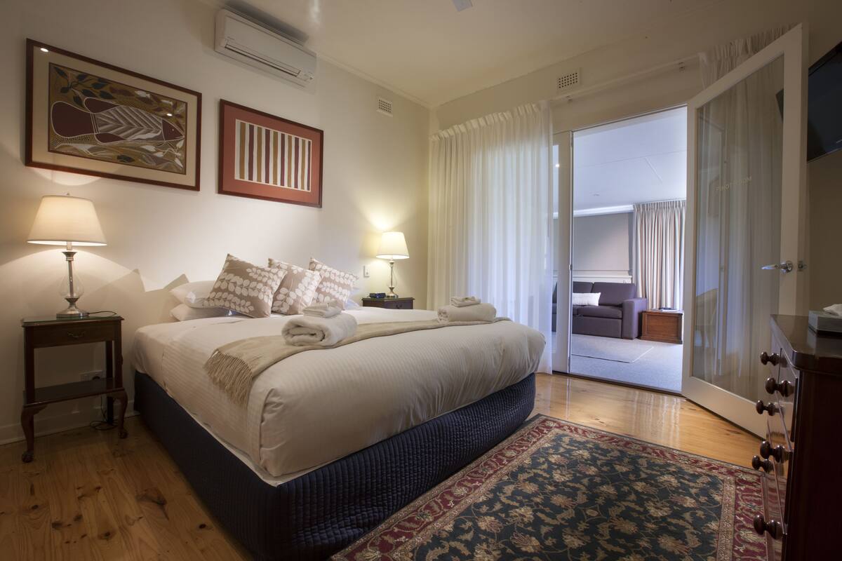 Un dormitorio con una cama grande, dos mesitas de noche con lámparas, una alfombra estampada y vistas a un balcón con sofá.