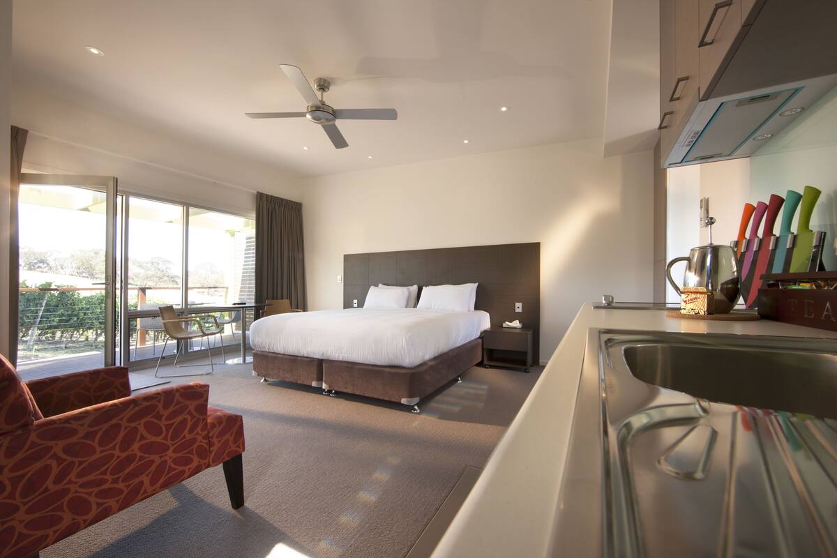 Una habitación de hotel moderna con una cama grande, ventilador de techo, balcón con una silla y una zona de cocina con utensilios.